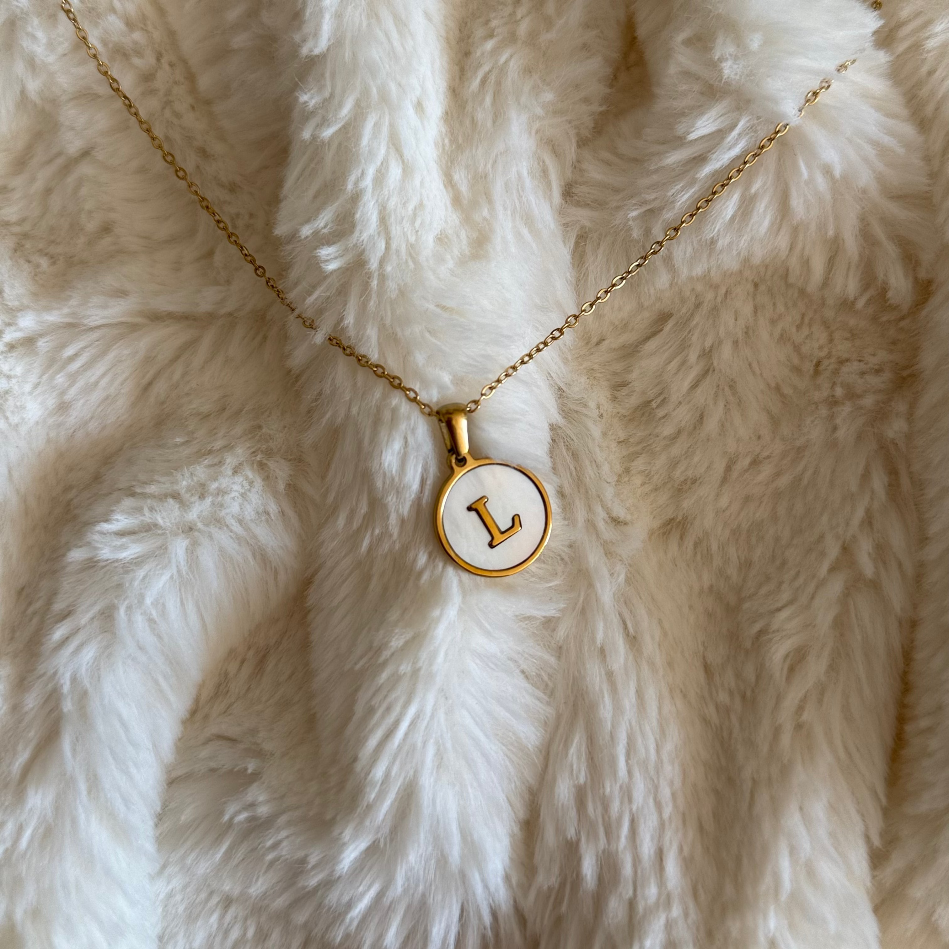 Gold necklace with a circular pendant featuring the letter 'L' on a fluffy white surface.
